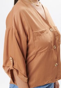 Blusa corta a maniche corte di color marrone chiaro, realizzata in tessuto morbido, con scollatura a V profonda, due tasche frontali e dettagli con bottoni sulle maniche.