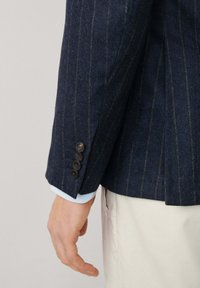 Námořnický modrý blazer s pruhy vyrobený z texturované látky, který má na rukávu čtyři černé knoflíky a pod ním je vidět světle modrá manžeta košile.