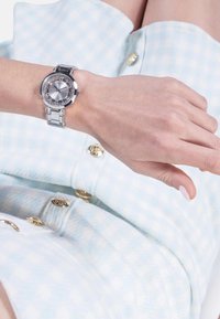 Hõbe käekell ümmarguse näo, kristallaktsentide, metallist rihma ja päikeseprismadialiga, kantud käe peal helesinise ruudulise kangaga.