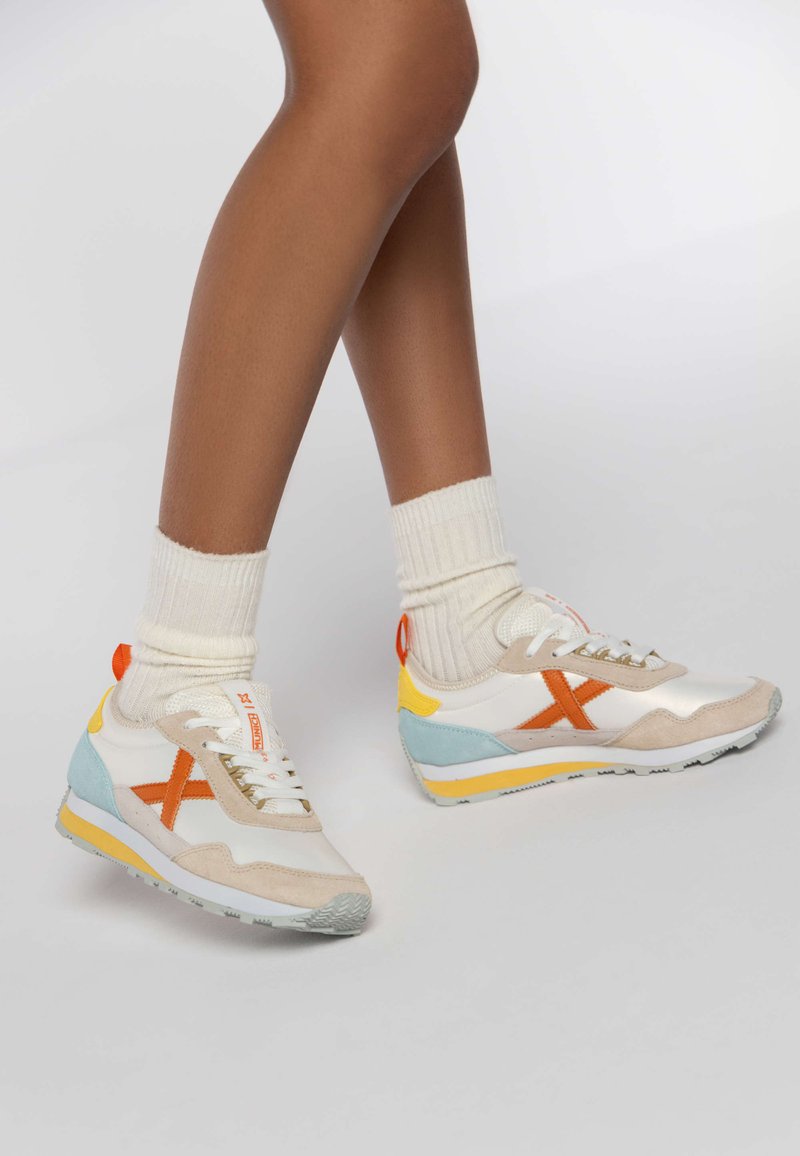 White and pastel sneakers with orange accents, suede and mesh materials, triangular blue and yellow details, and textured soles.