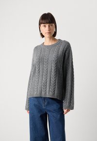 Pull en tricot gris avec un col rond et des manches longues, doté d'une finition côtelée et d'un motif texturé. Associé à un jean bleu à jambes larges.