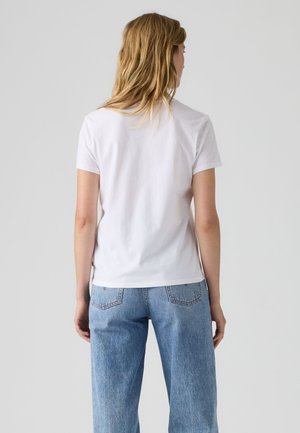 Mujer con cabello rubio que lleva una camiseta blanca lisa de manga corta y vaqueros azules, de pie de espaldas contra un fondo liso.