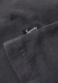 Ciemnoszara tkanina o miękkiej teksturze. Posiada widoczną kieszeń oraz zieloną metkę z logo "Superdry" przyszytą do materiału.