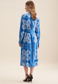 Abito blu a maniche lunghe con intricati motivi a paisley bianchi, vita aderente e gonna di media lunghezza. Indossato con scarpe con tacco nere.