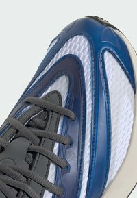 Sneaker with a textured mesh upper in white, accented by smooth blue overlays and gray laces, featuring a flexible design and visible stitching.