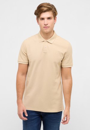 Junger Mann trägt ein beigefarbenes Polohemd mit kurzen Ärmeln und blaue Jeans, steht vor einem schlichten hellen Hintergrund.