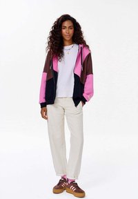 Rozpinana bluza z kolorowymi blokami w odcieniach różu, brązu i czerni, zestawiona z białym t-shirtem i kremowymi spodniami. Brązowe sneakersy z różowymi akcentami.