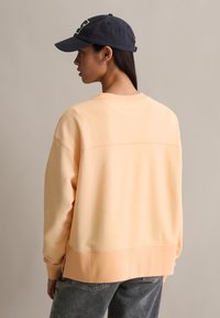 Sweat-shirt pêche avec une surface texturée et des poignets côtelés, associé à une casquette marine avec un logo contrastant, et un jean gris décontracté.