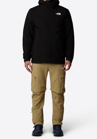 Schwarze wasserdichte Jacke mit Kapuze, kombiniert mit beigen abnehmbaren Hosen mit Cargo-Taschen. Dazu werden blaue Wanderschuhe getragen.