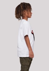 Weißes Baumwoll-T-Shirt mit grafischem Druck, übergroßer Schnitt, kurze Ärmel. Getragen mit olivgrünen Cargo-Hosen. Steht vor einem neutralen Hintergrund.