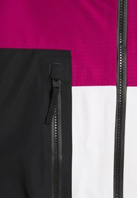 Detailní záběr na tříbarevnou bundu s černou zipovou kapsou a vertikálním zipem, která obsahuje panelové části v magentové, bílé a černé barvě.