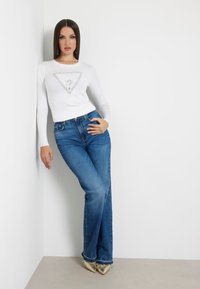 Maglietta bianca a maniche lunghe con un'applicazione a forma di diamante, abbinata a jeans a zampa d'elefante blu e scarpe metalliche a punta. Sfondo minimalista.