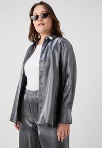 Metallic silver button-up shirt with long sleeves, smooth texture, and black sunglasses, paired with matching shiny pants and a white top.