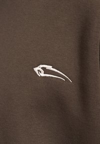 Top in tessuto marrone con superficie testurizzata e logo ricamato bianco sul lato sinistro, che aggiunge contrasto al design complessivo.