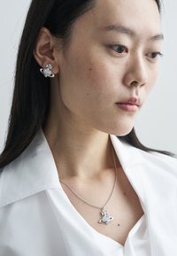 Strieborný prívesok na náhrdelníku s motívom planéty, doplnený o zodpovedajúce kruhové náušnice. Nosené na bielom tričku, s lesklým povrchom.