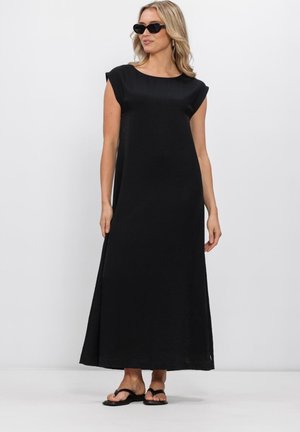 Vrouw met zwarte mouwloze maxijurk, zwarte zonnebril en zwarte platte sandalen, staand tegen een effen witte achtergrond.