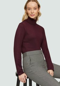 Bordeaux coltrui in ribbelstructuur, gecombineerd met grijze broek met een gestructureerde tailleband en zijzakken, zittend op een kruk.