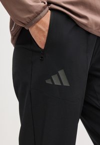 Main dans la poche d’un pantalon de sport noir avec poche zippée et logo Adidas, porté avec un haut marron à manches longues.
