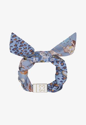 Coletero de tela azul con estampados florales y de leopardo, con un lazo y un cierre rectangular dorado y blanco con diseño de trébol.