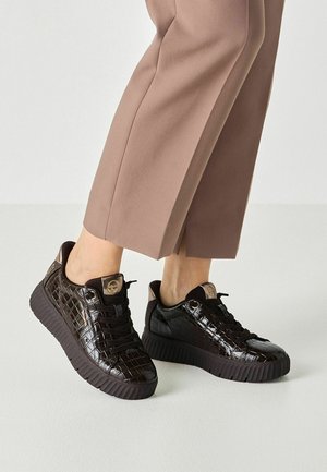 Skinnende sorte sneakers i krokodillemønster med tykke såler, båret med lysebrune korte bukser på en neutral baggrund.