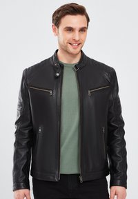 Zwarte leren jas met ribdetails op de schouders, ritszakken en een iets getailleerd ontwerp. Gelaagd over een groene shirt.