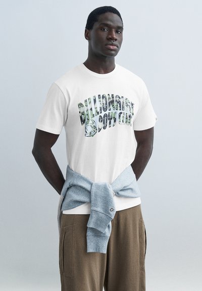 Young man wearing white Billionaire Boys Club t-shirt, light gray sweatshirt tied around waist, and brown pants against a plain gray background.