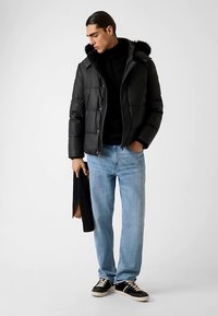Schwarze Steppjacke mit Kapuze, die mit Fell besetzt ist, getragen über einem schwarzen Pullover. Hellblaue, gerade geschnittene Jeans und schwarze Turnschuhe vervollständigen das Outfit.