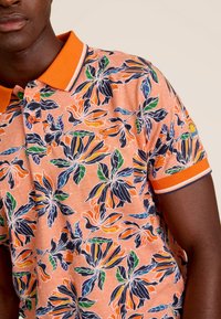 Korte mouwen polo shirt in oranje met een bloemmotief. Beschikt over een kraag en boorden met contrasterende oranje en marineblauwe afwerking. Knopen aan de voorkant.