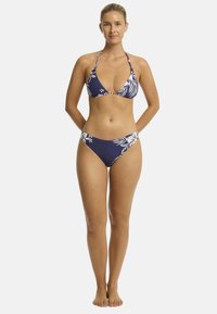 Modelo en un bikini floral azul marino de pie, con las manos detrás de la espalda y sonriendo. El fondo neutro resalta el diseño del traje de baño.