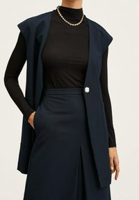 Blazer senza maniche blu navy con chiusura a bottone singolo, indossato sopra una maglietta a dolcevita nera, caratterizzato da una texture liscia e un design su misura.
