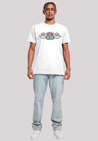 White cotton t-shirt featuring a colorful "CENTRAL PERK" graphic, short sleeves, worn with light blue denim jeans and white sneakers.
