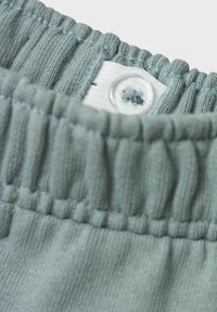 Světle zelený žebrovaný pásek s knoflíkovým zapínáním a bílou podšívkou. Texturovaná plocha vykazuje měkký, elastický materiál.