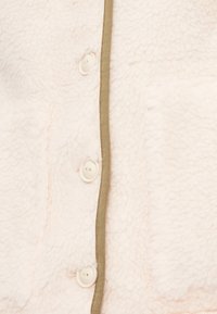 Vêtement d'extérieur en polaire de couleur rose doux, présentant une surface texturée, un biais kaki le long du bord et des boutons clairs pour la fermeture.