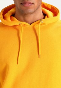 Felpa con cappuccio gialla brillante in cotone, dotata di cappuccio regolabile con cordino, occhielli in metallo e superficie di tessuto testurizzato.