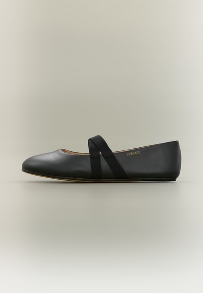 Chaussure plate en cuir noir avec des brides élastiques croisées et un petit talon, estampillée du petit logo Versace doré sur le côté.