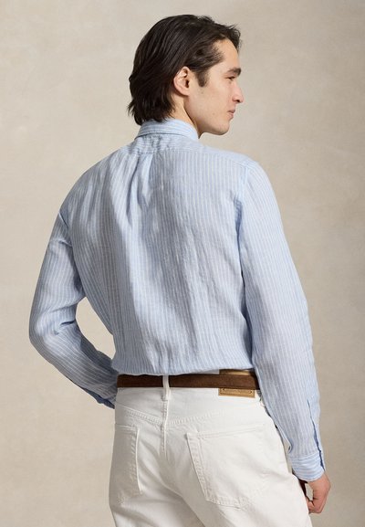 Mees seljaga vastu, kandes helesinist triibulist nööbitavat särki, mis on valgete pükstega ja pruuni vööga kokku pistetud, ühevärvilise tausta ees.