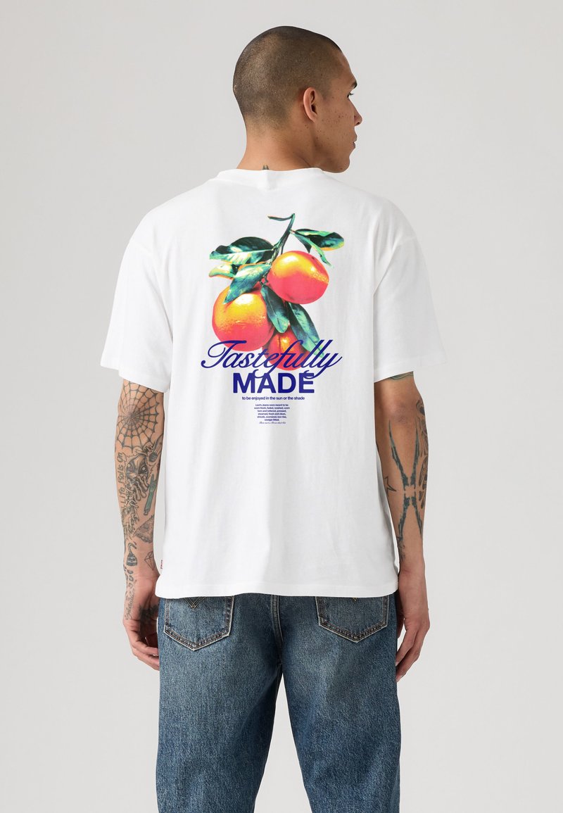 Άνδρας με τατουάζ φοράει λευκό T-shirt που δείχνει τύπωμα στην πλάτη με πορτοκάλια σε κλαδί και το κείμενο «Tastefully MADE» πάνω από μικρό κείμενο.