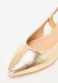 Chaussure plate slingback en métal doré, avec un bout pointu, une surface texturée et un design découpé. L'intérieur présente un revêtement lisse beige.