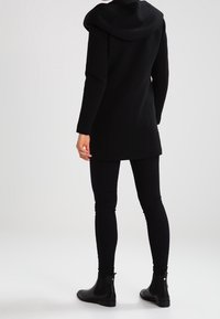 Abrigo negro con capucha drapeada, corte recto, tejido texturizado, llevado sobre leggins negros ajustados, combinado con botines negros.