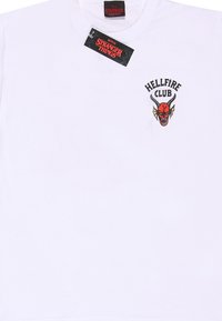 Bílá bavlněná tričko s barevným grafickým designem "Hellfire Club" s démonem s rohy na levé části hrudi, s připojenou etiketou.