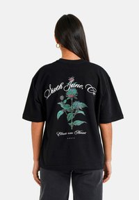 Camiseta negra de gran tamaño con un gráfico floral en verdes y azules, con texto blanco que dice "Sixth June, Co" y "Eleva cada Momento" abajo.