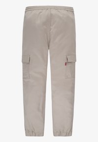 Beżowe spodnie cargo z elastycznym pasem i mankietami, z klapowymi kieszeniami na obu udach oraz małą czerwoną metką na prawej kieszeni.