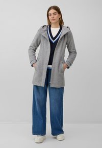 Manteau à capuche gris avec une fermeture éclair à l'avant et deux poches latérales, superposé sur un pull bleu marine à col en V avec des accents blancs. Pantalons en denim bleu larges.
