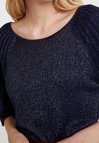 Marineblauer Pullover mit funkelnder Textur, rundem Ausschnitt und plissierten Details an den Schultern. Weiches Strickmaterial.