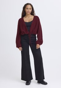 Maglione corto bordeaux con davanti aperto, dettaglio con coulisse e maniche lunghe, abbinato a pantaloni neri a gamba larga e stivaletti alla caviglia.