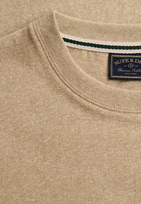 Pull beige à col rond avec col côtelé et étiquette bleu marine indiquant "Superdry Premium Outfitters New York 1954" cousue à l'intérieur de l'encolure.
