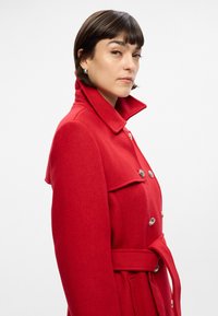 Rode trenchcoat van textuurwol, met een hoge kraag, een taille met riem en zilveren knopen aan de voorkant. Soepele, getailleerde silhoutte.