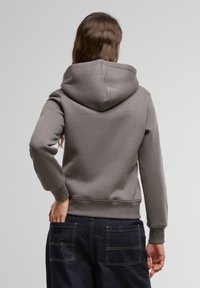 Sweatshirt gris avec une capuche spacieuse, des poignets et un ourlet côtelés, associé à un pantalon en denim foncé avec des poches arrière doubles. Texture lisse.