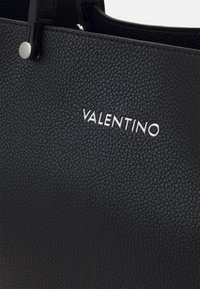 Närbild av en svart texturerad läderhandväska med silverfärgad Valentino-logotyp och ett handtag som säkrats med en silverknapp.