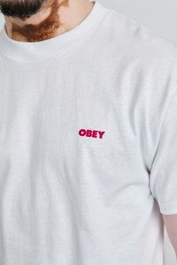 Camiseta de algodão branca com um pequeno logótipo vermelho "OBEY" no lado esquerdo do peito. Mangas curtas e gola redonda com uma textura suave.